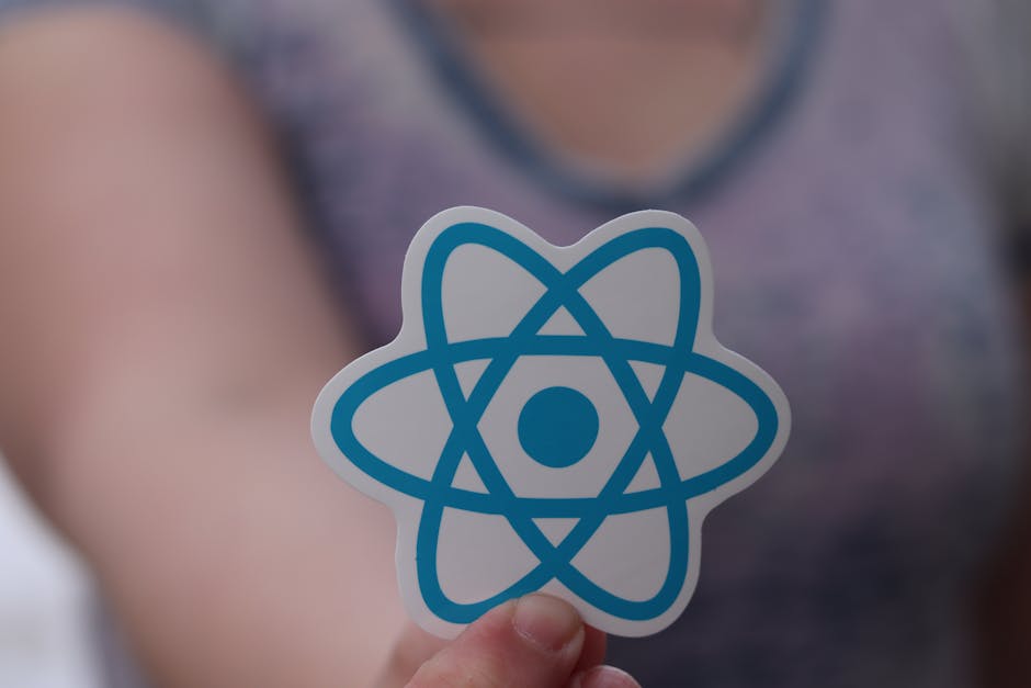 Close-up of a person holding a sticker featuring a blue atom symbol, with the blurred background of a person wearing a grey tank top and visible skin tone, indoors under soft lighting. The image is as